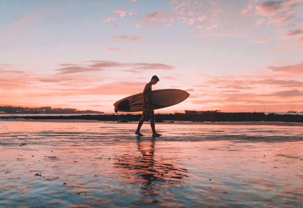 hombre sujetando la tabla de surf caminando cerca de la orilla del mar rompecabezas en línea