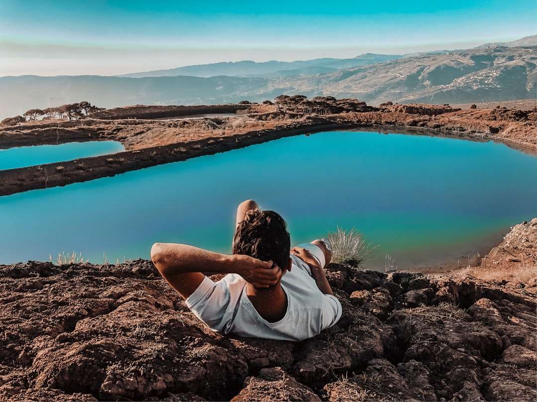 donna in camicia bianca che si siede sulla roccia marrone vicino al lago puzzle online