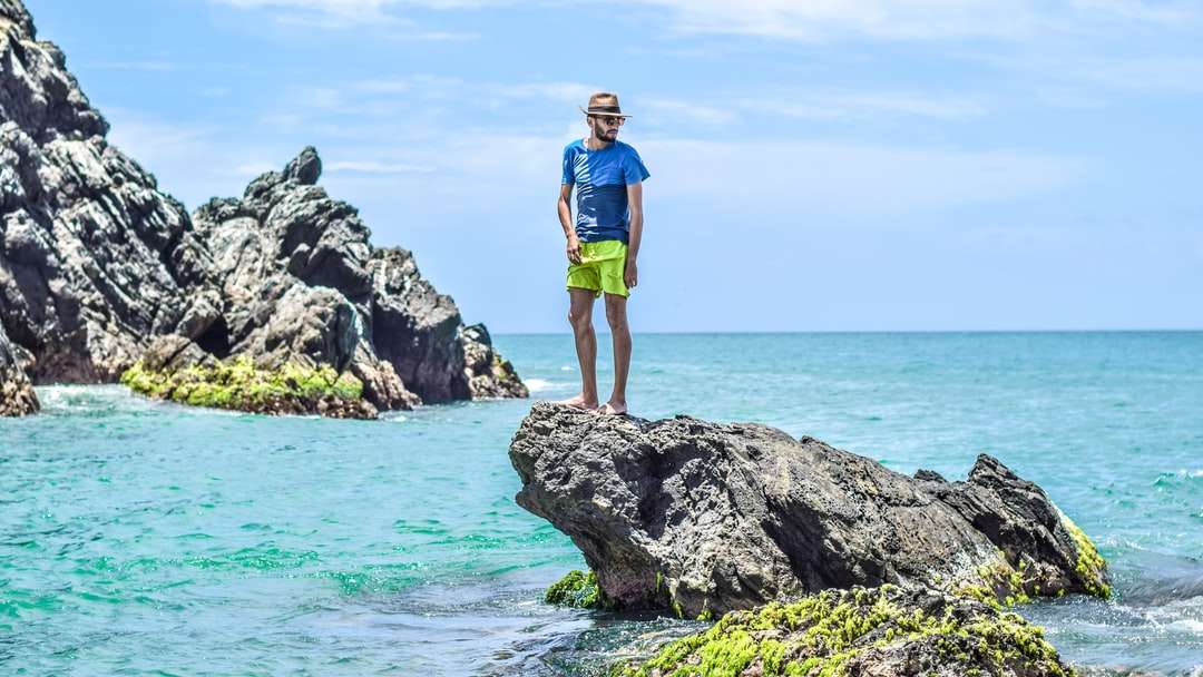 Hombre con camiseta azul de pie sobre una roca gris en el mar rompecabezas en línea