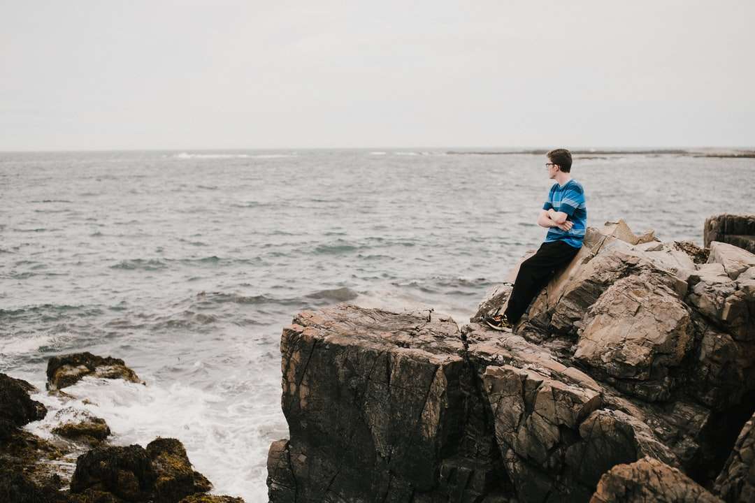 Mann im blauen Hemd, das auf Felsformation nahe Meer sitzt Puzzlespiel online