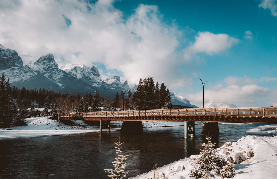 καφέ ξύλινη γέφυρα πάνω από τον ποταμό κοντά στο χιονισμένο βουνό online παζλ