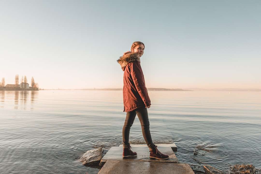 vrouw in rode jas staande op rots in de buurt van zee overdag online puzzel