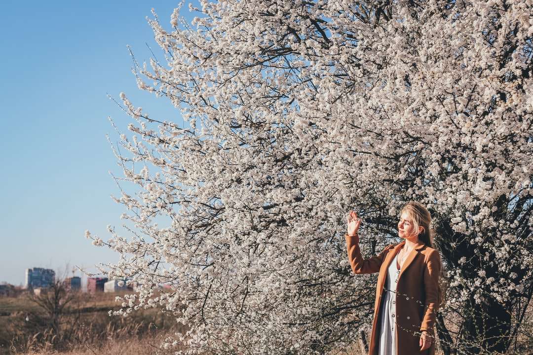 woman in brown coat standing near white cherry blossom tree online puzzle