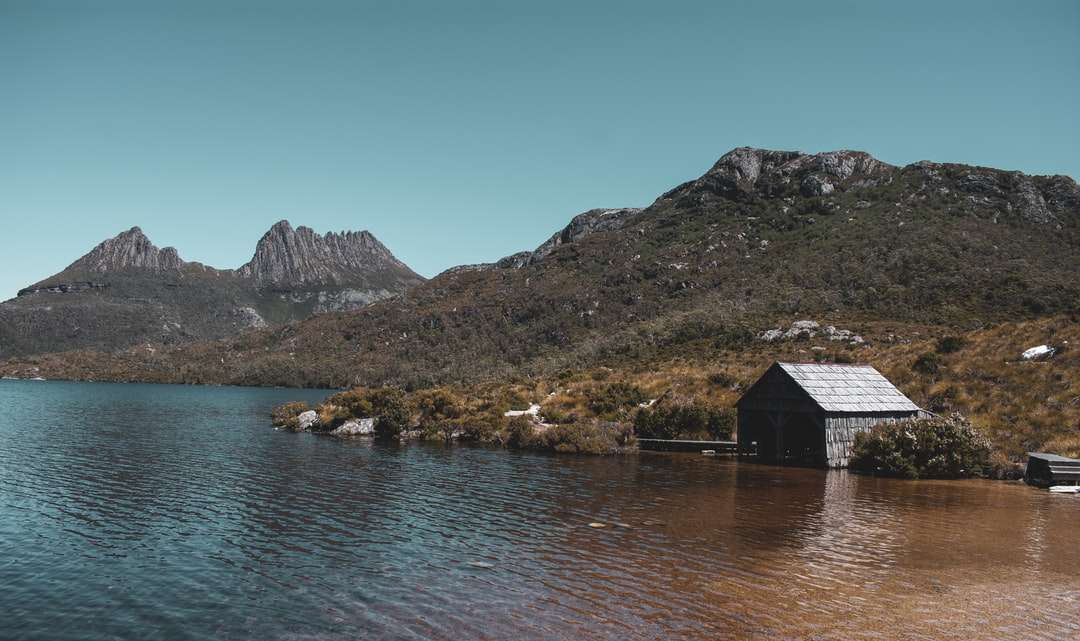 Καφέ ξύλινο σπίτι κοντά στη λίμνη και το βουνό κάτω από τον μπλε ουρανό παζλ online