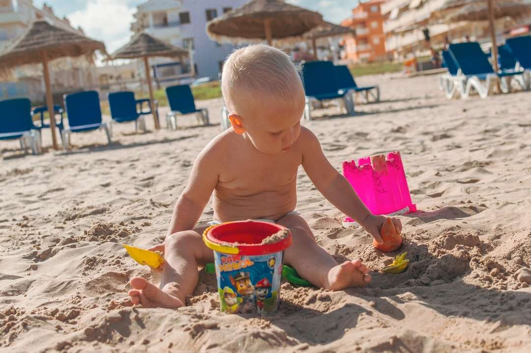 Niño en topless con cubo de plástico azul en la arena rompecabezas en línea