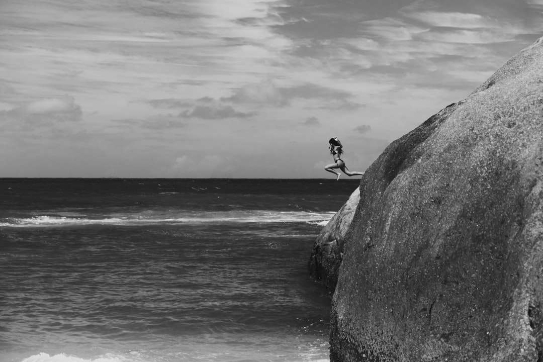 Foto en escala de grises del hombre sentado en la roca cerca del mar rompecabezas en línea