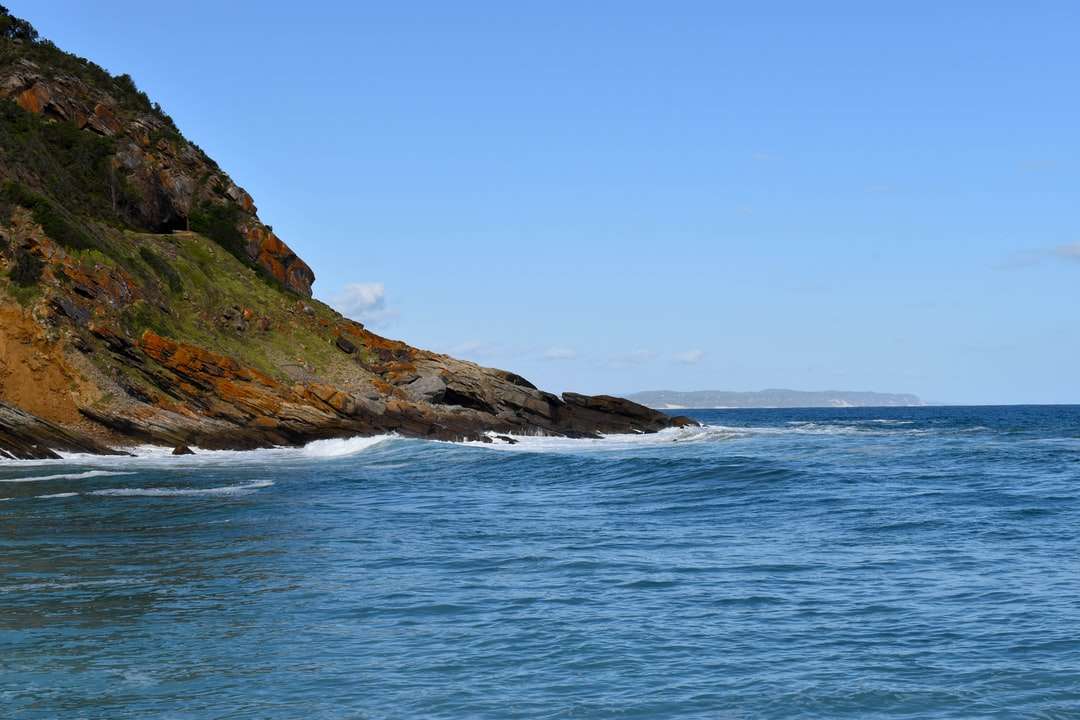 Formación de roca marrón y verde al lado del mar azul. rompecabezas en línea