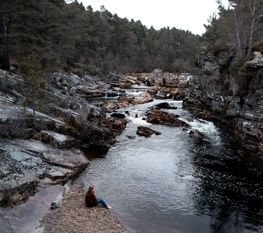 Persona en chaqueta azul sentado en roca cerca del río rompecabezas en línea