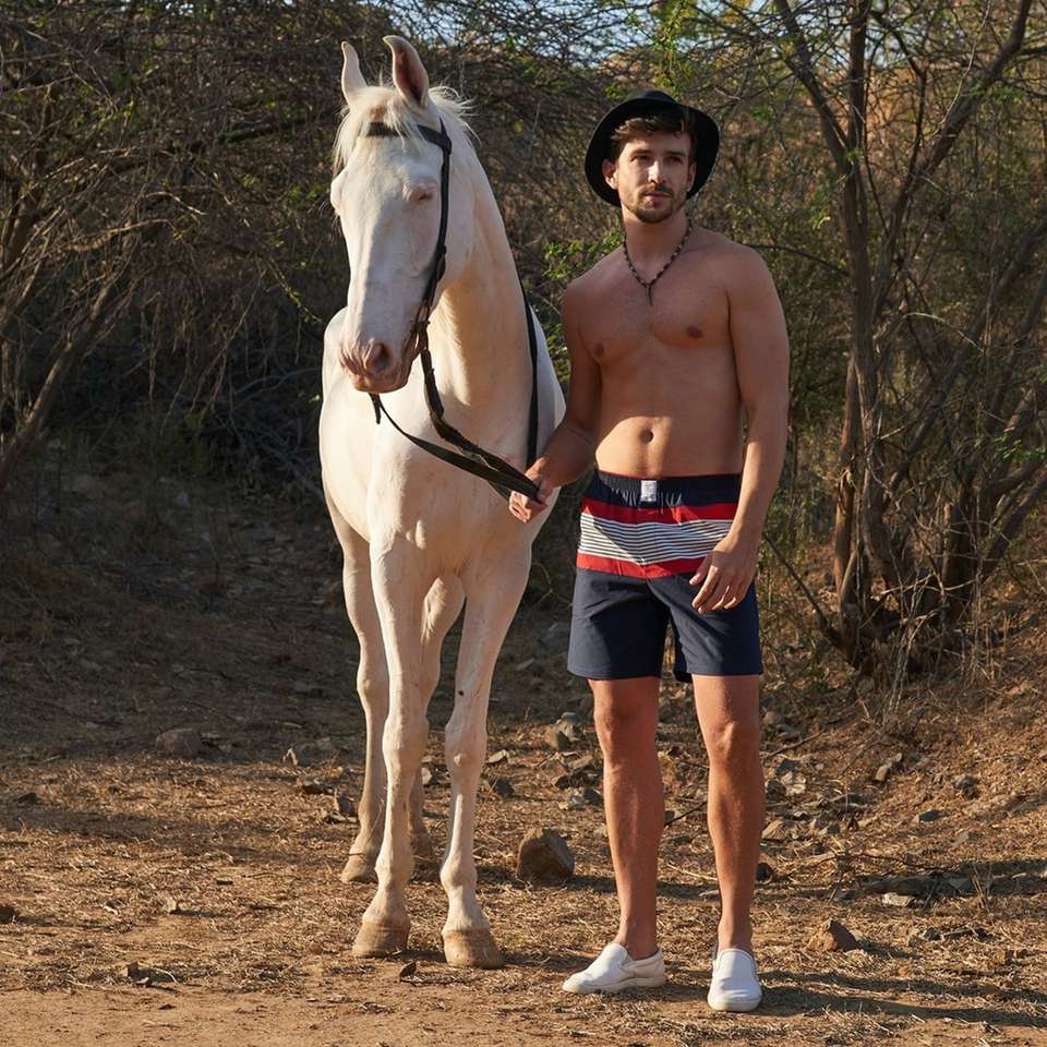 Omul topless în pantaloni scurți albastri stând lângă calul alb puzzle online