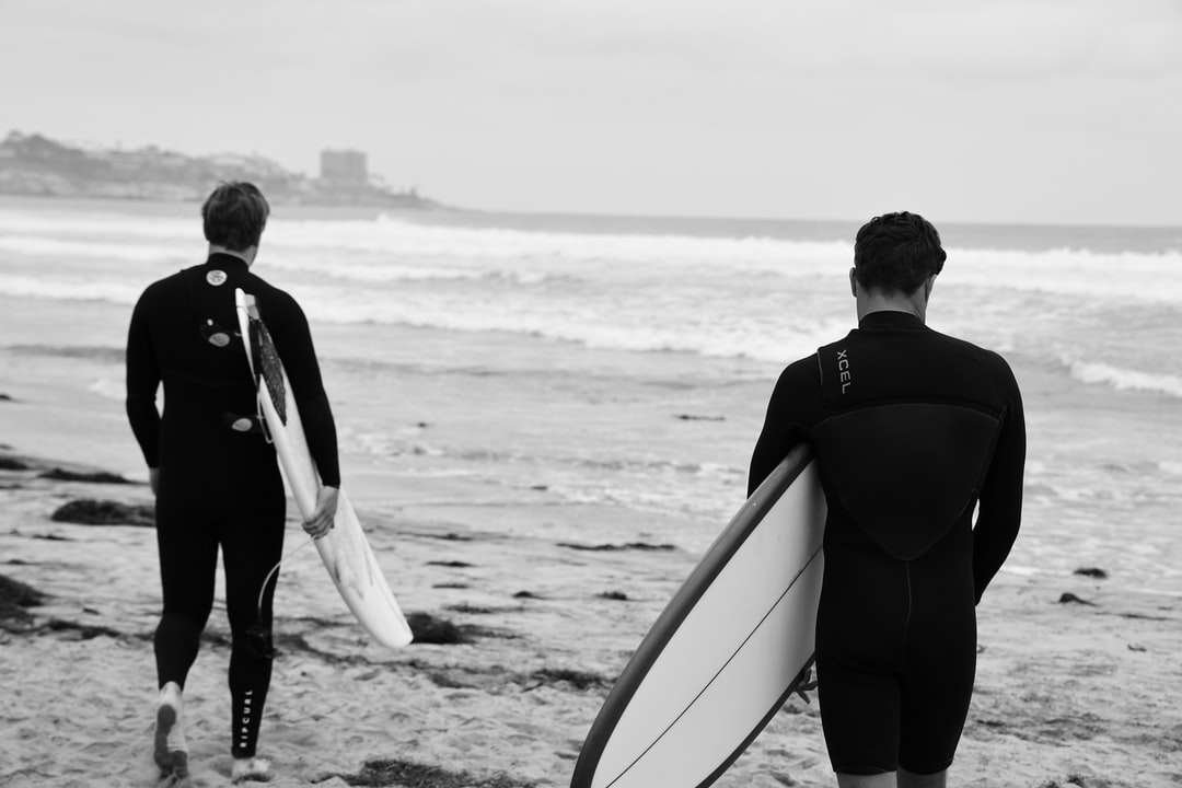 мужчина в черной куртке с белой доской для серфинга идет по пляжу онлайн-пазл