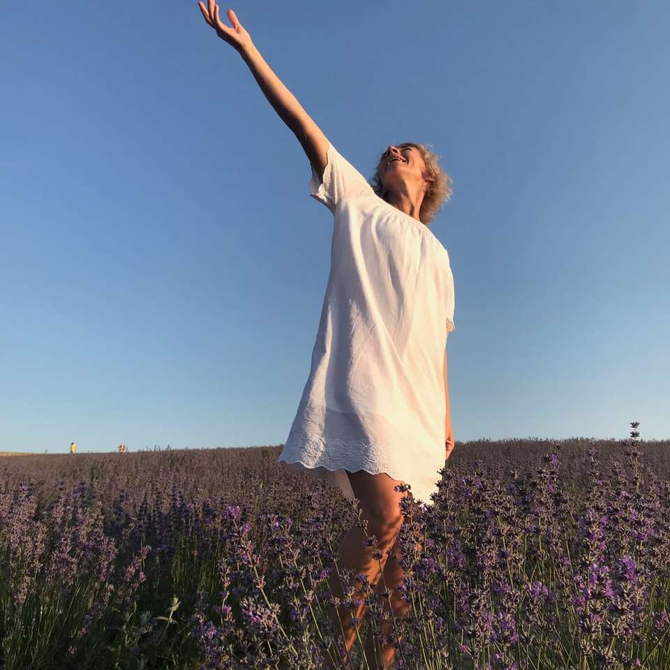 жена в бяла риза, стояща на пурпурно поле на цветя онлайн пъзел