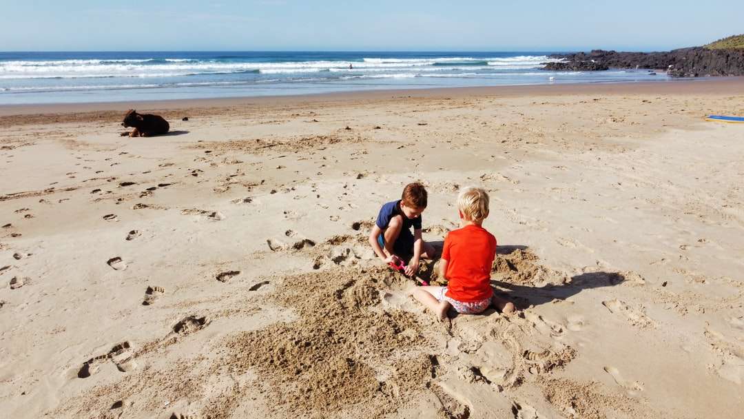 2 niños sentados en arena marrón cerca del mar durante el día rompecabezas en línea