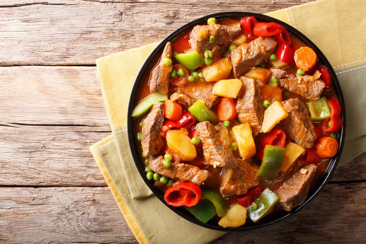 Ázsiai étel: Párolt marhahús burgonyával, paprikával, borsóval, paradicsommal és sárgarépával közelről egy tányéron az asztalon. online puzzle
