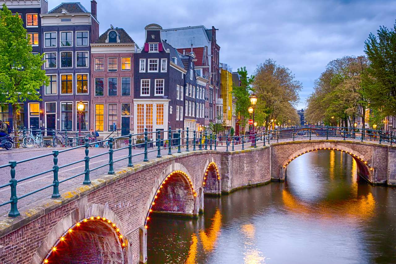 Éjszakai kilátás Amszterdam városképe a csatornákkal. Megvilágított híd és hagyományos holland házak szürkületben a háttérben. Vízszintes lövés online puzzle